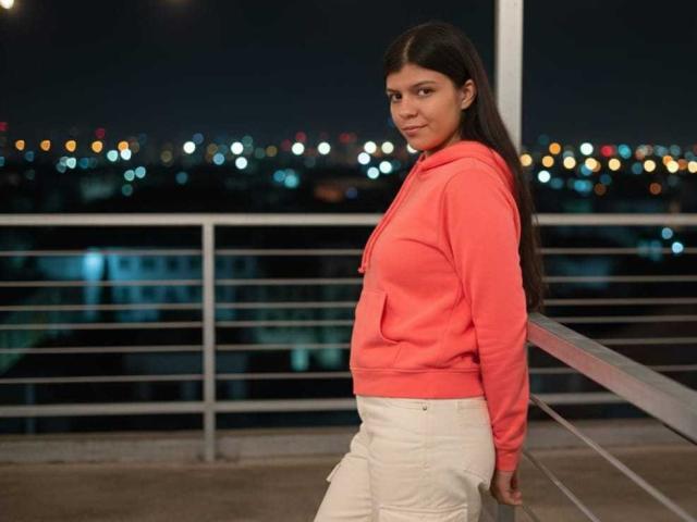 RilleyFoyx stands with a slight smile, wearing a pink hoodie and white pants, her ass slightly visible, against a metal railing at night with city lights in the background.