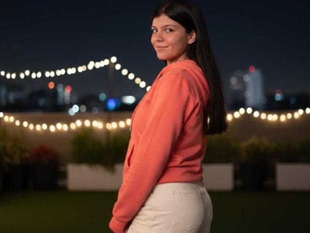RilleyFoyx with long black hair, wearing a pink hoodie and white pants, smiling at the camera, standing on a rooftop at night with string lights in the background.