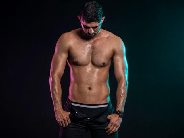 TonnyRice is a muscular, shirtless dude with a sweaty, glistening chest, dark pants, and a watch, looking down with his hands in his pants.