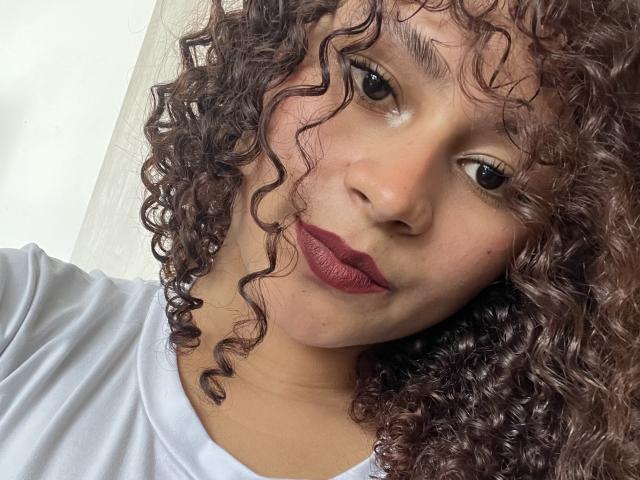 SofiaPassionne with curly brown hair, medium skin, red lipstick, and a white shirt, looking at the camera with a slight smile. Soft natural light highlights her face.