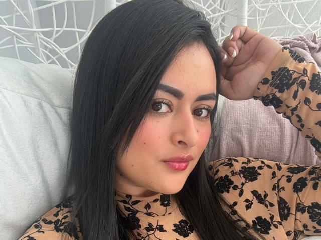 AmberGamble has dark, straight hair, wearing a floral beige top, with pink lipstick, and a slight smile, looking at the camera with her hand in her hair.