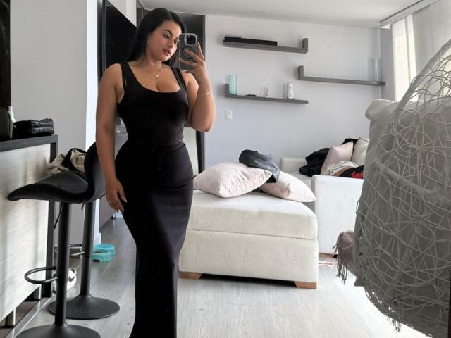 AmberGamble, curvy, large tits, wearing a tight black dress, taking a mirror selfie, standing in a modern living room, holding a phone, with a chair to the left.