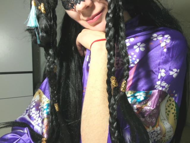 AromaLove with long black braids, wearing a purple floral kimono, black lace mask, and a red bracelet, smiling with a hand on their chin, in a well-lit room.
