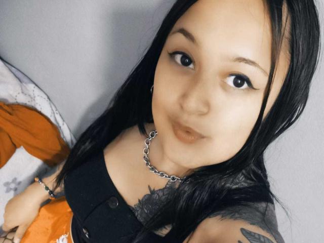 MeganQuenn with long black hair, wearing a black top with buttons, silver chain necklace, and tattoo on her right shoulder, looking up with pouty lips.