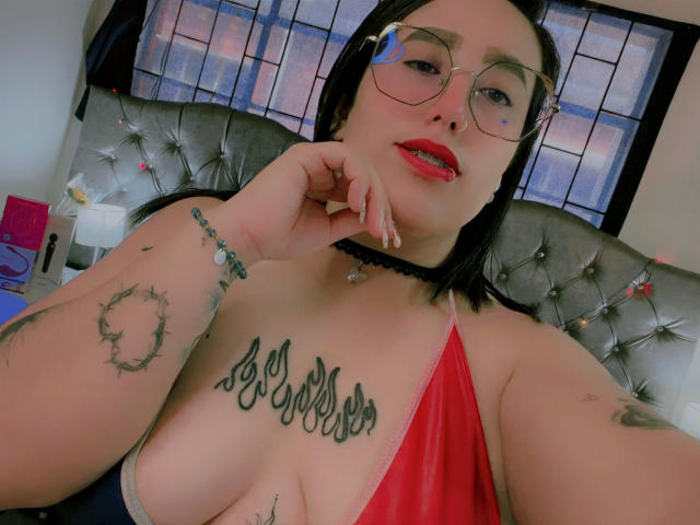 NatalieHarris with glasses, red lipstick, black choker, tattoos on chest and arm, red halter top, hand on chin, looking at camera, bedroom, windows in background.