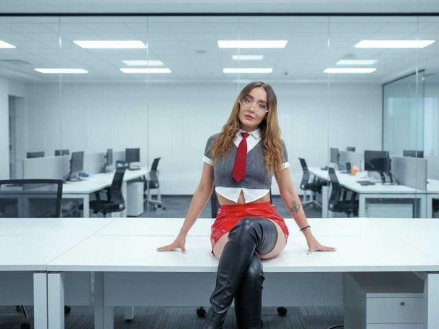 MindyLove sits on a white office desk, wearing a grey crop top, red tie, red mini skirt, and black thigh-high boots. She has long brown hair and tattoos.