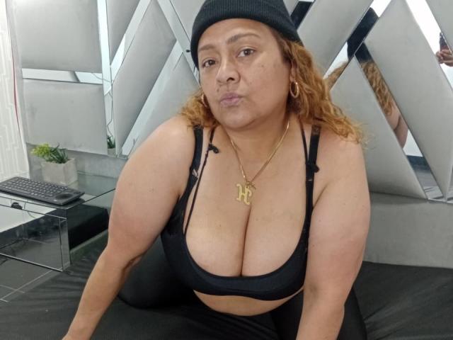 Monzerrat Boyer, con i capelli arancioni ricci, un berretto nero, un reggiseno sportivo nero e una collana dorata, con le labbra tumefatte, che mostra il suo seno prosperoso, seduta su un letto nero, mentre guarda la fotocamera..