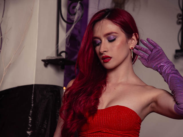 AndraCyrus with red hair, purple gloves, red strapless dress, purple eyeshadow, red lipstick, and earrings, touching her ear, showing her bare shoulders and small tits.
