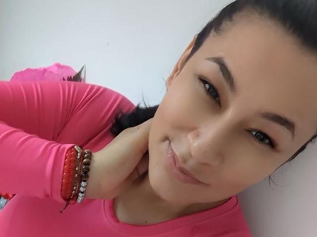 EmaFoxy has light brown skin, black hair in a ponytail, wearing a pink long-sleeve top, red and brown bracelets, and a slight smile with her hand near her neck.