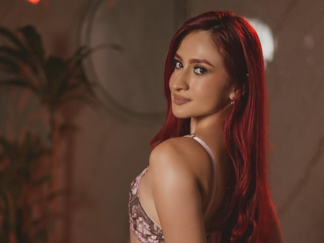 AndraCyrus with red hair, looking over her shoulder, wearing a pink lace bra, has small tits, a cute face, and a subtle smile. Soft lighting, warm tone.