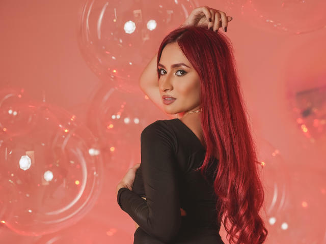 AndraCyrus with long red hair, black top, looking over shoulder, hand in hair, glossy lips, subtle makeup, soft red lighting, clear balloons in background.