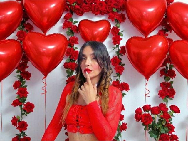 MindyLove mit rotem Lippenstift, Finger am Mund, trägt ein rotes, durchsichtiges Oberteil mit langen Ärmeln und ist umgeben von roten Herzballons und Rosen. Ihre Brüste sind vom Oberteil bedeckt..