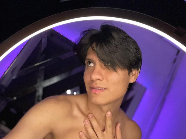 Shirtless AlexxNorth with short black hair, medium skin, and a cute face, hand on chest, looking slightly to the side, purple ring light in background.