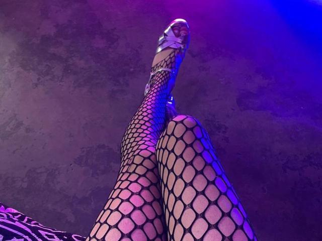 AlexxNorth's legs in black fishnet stockings, pointed high heels, and shiny silver ankle straps, shot from a low angle with purple and blue lighting.