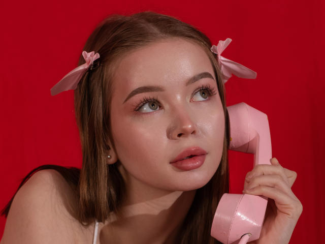 ViolettaFane with pink hair bows, holding a pink phone against a red background, has shiny skin and full lips, looking to the side with a subtle smile.