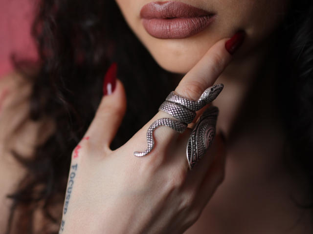 Venusinylons' vollschlanke Lippen, dunkles, welliges Haar, roter Nagellack, silberner Schlangenring, tätowierter Finger, Ring in der Nähe des Mundes. Brüste teilweise sichtbar, zarter rosa Lippenstift..