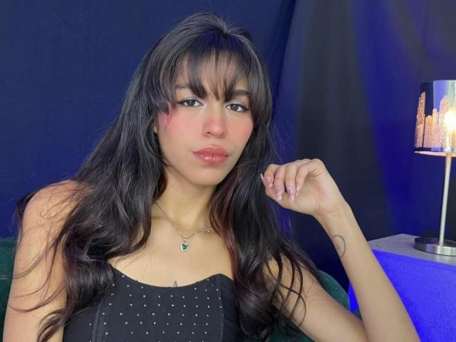 SoffiaCollins has long black hair, bangs, and wears a black polka dot top. She's looking at the camera with pink lips and a small tattoo on her right wrist.