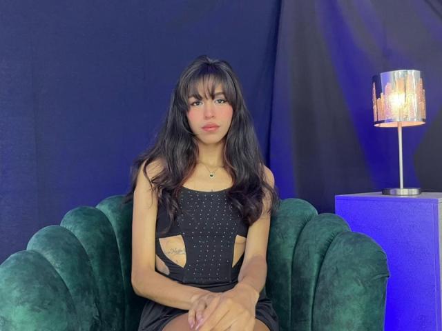 SoffiaCollins with long black hair, wearing a black cutout dress, sitting on a green velvet chair, looking at the camera, hands on lap, small tattoo on left arm.