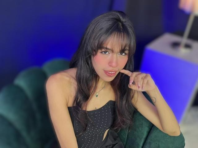 SoffiaCollins with long black hair, pink blush on cheeks, wearing a black strapless top with small white dots, finger to mouth, green velvet chair, blue background.