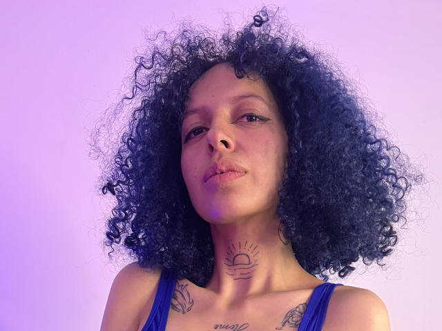 HannahBlue with curly black hair, blue tank top, tattoos on neck and chest, pouty lips, and a subtle purple light on her right side. Her tits are small.
