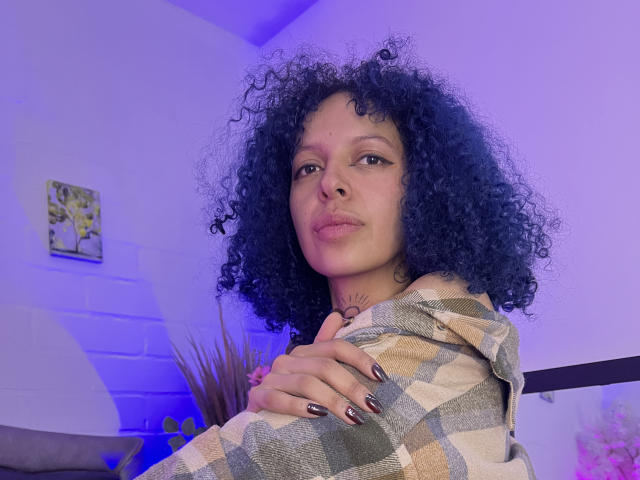 HannahBlue with curly black hair, light brown skin, and dark eyes, wearing a plaid shirt, puffs her pink lips, and holds her arm, showing black and silver nails.