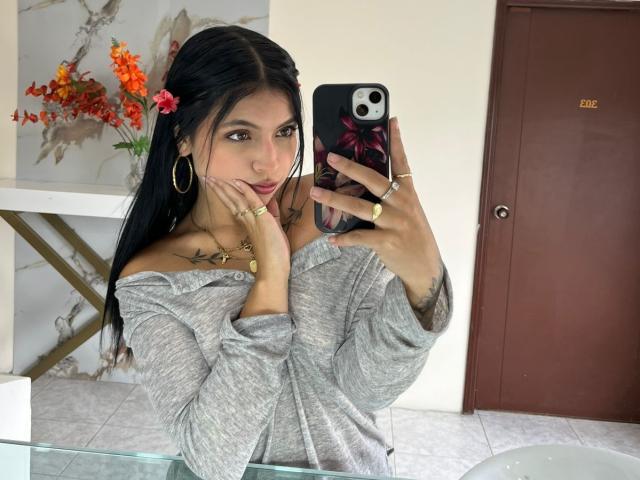 NyraVanel with long black hair, gray off-shoulder top, gold hoop earrings, and rings, taking a mirror selfie with her phone, showing tattoos and a pink flower in her hair.