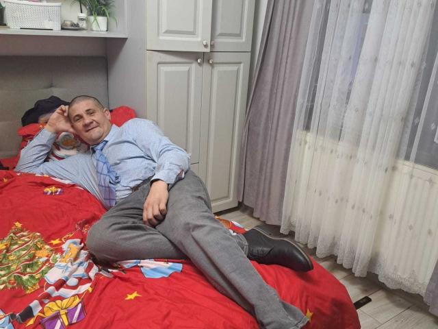 CrisSalon, a middle-aged man with a shaved head, is lying on a red bed with colorful patterns. He wears a light blue shirt, gray pants, and black shoes. His right arm is bent with his hand resting on his forehead, and his left arm is relaxed. The bed has bright red sheets with yellow stars and a small blue pillow. White curtains and a white cabinet are in the background. The room is well-lit with natural light.