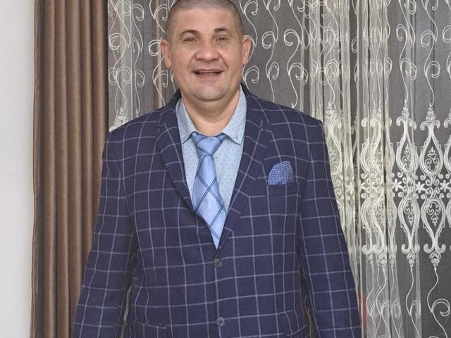 CrisSalon, a middle-aged man with a shaved head and light skin, stands in a formal setting. He wears a dark blue, plaid-patterned suit with a light blue shirt and a matching tie. A blue pocket square is in the suit's left chest pocket. The background features ornate, white lace curtains with brown vertical panels on the left. CrisSalon's relaxed, slightly smiling expression is visible. The image focuses on his attire and upper body.