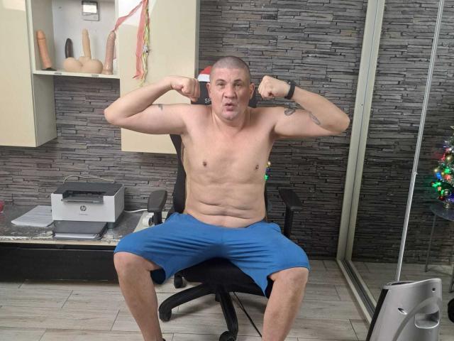 CrisSalon, a shirtless man with a bald head and tattoos on his arms, sits in a black office chair with legs spread. He's flexing his biceps, wearing blue shorts, and has a surprised expression. The room has a gray stone wall, a shelf with sex toys, and a printer on a desk. A black wristband is on his right wrist. The floor is tiled. The image is a photograph.