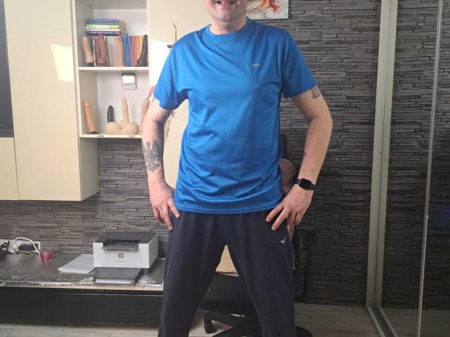 CrisSalon stands in a room wearing a bright blue t-shirt, black athletic pants, and a black smartwatch. CrisSalon's mouth is open in a playful expression, hands on hips. Tattoos cover both arms. Background includes a shelf with books and sex toys, a printer on a desk, and a gray stone-textured wall. CrisSalon is centered, with the room's details slightly blurred.