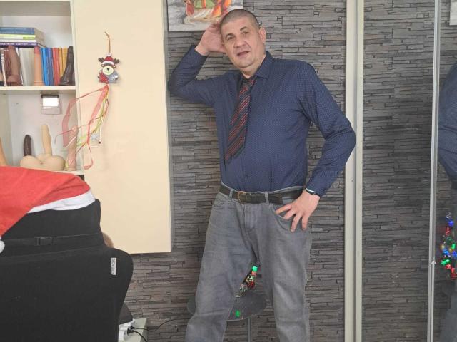 CrisSalon, bald, blue shirt, striped tie, grey pants, belt, standing, hand on hip, other hand on head, smiling, bookshelf, wall, mirror, colorful decoration, chair with red blanket.