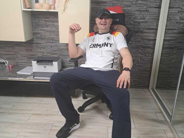 CrisSalon, a chubby dude with a German shirt, black cap, blue pants, and black shoes, sitting on a chair, flexing his right arm, smiling, in an office.