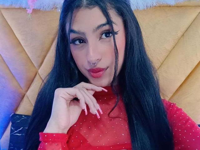 LilithDesire with long black hair, red sheer top, pink lipstick, septum piercing, eyeliner, and pink nails, looking at camera, hand near face, sexy, confident.