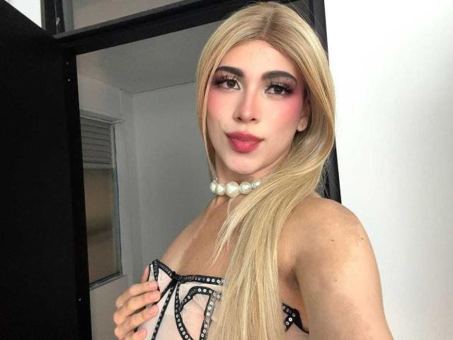 LyaCollins with long blonde hair, pink eyeshadow, and a pearl necklace, wearing a black strapless dress with white studs, covering her tits with one hand.