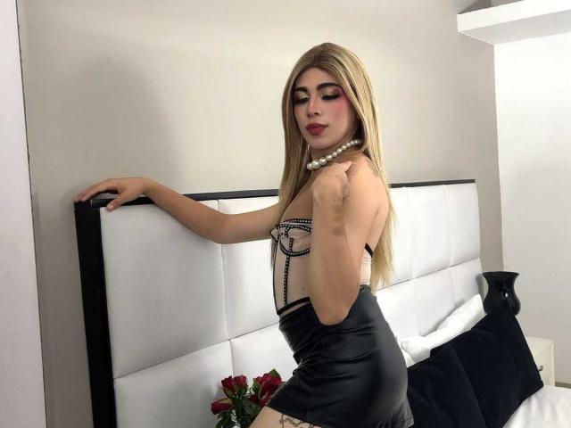 LyaCollins with long blonde hair, wearing a black leather mini-dress, pearl necklace, and chain lingerie, leaning on a white and black headboard, showing her small tits and ass.