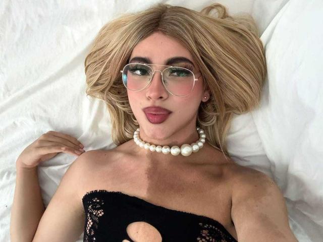 LyaCollins with blonde hair, glasses, and pearl necklace, lying on white bed, wearing black lace top with cutout, showing small tits, pouting with pink lips, right hand near head.