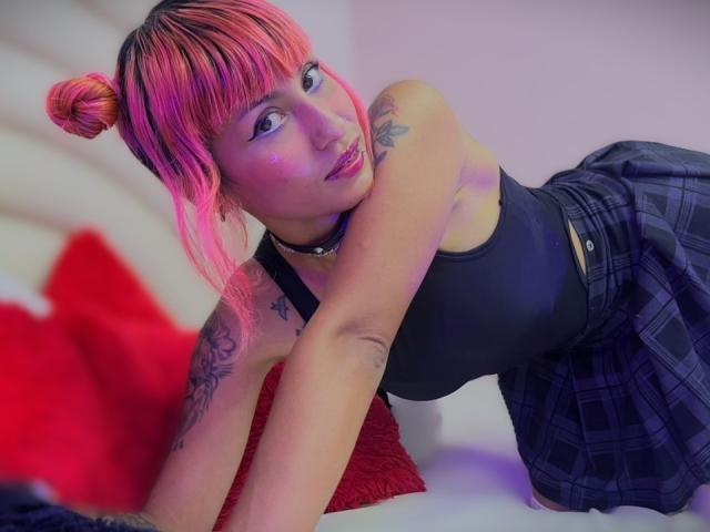 MilaRouge with pink and orange hair in a bun, black tank top, plaid skirt, tattoos, and a choker, leaning on a red pillow, looking at the camera.