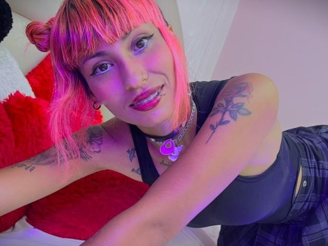 MilaRouge with pink hair, black tank top, plaid skirt, tattoos, heart necklace, and lip ring, smiling at the camera. Boob tattoos visible. Pink and purple lighting.