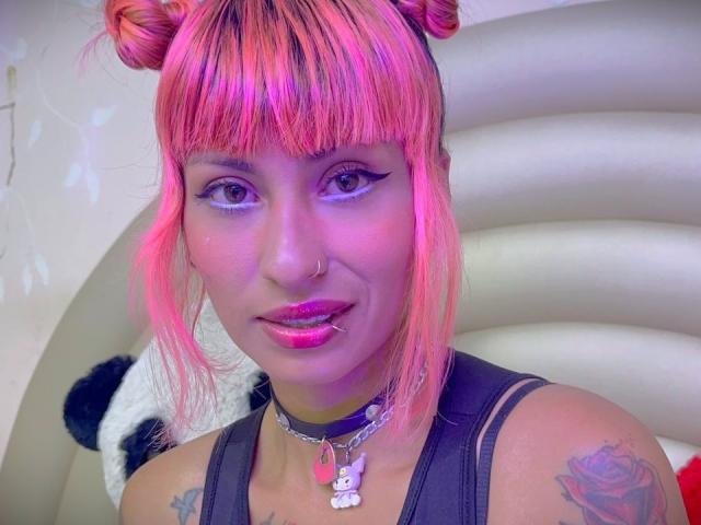 MilaRouge with pink hair in double buns, purple eyeliner, pink lipstick, nose ring, black tank top, choker with bunny pendant, tattoos, and a cute, sexy expression.