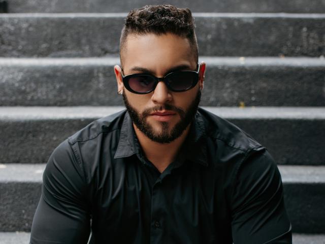 MassimoFiore has a beefy beard, dark sunglasses, and a black shirt. He's sitting on gray steps with a serious, slightly smug look. He's a chunky dude with curly hair.
