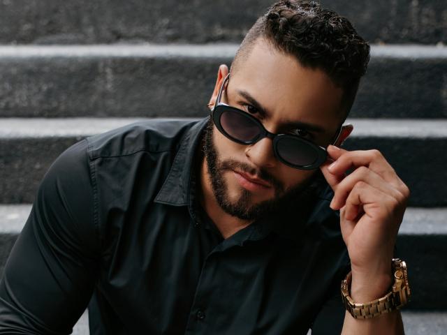 MassimoFiore with short curly hair, beard, black shirt, holding sunglasses, looking at camera, slightly smiling, gold wristwatch, dark skin, confident, sexy, handsome.