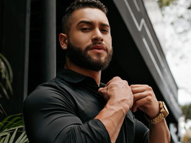 MassimoFiore with a dark beard, short curly hair, black shirt, gold watch, ear piercings, muscular arms, confident expression, leaning against a black structure, daytime, outdoors.