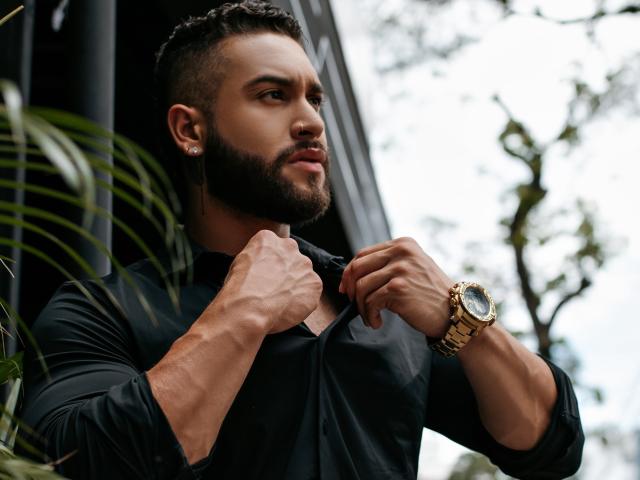 MassimoFiore, muscular, bearded man with dark skin, buttoning black shirt, gold watch, short curly hair, earrings, looking side, green plant in foreground, overcast sky in background.