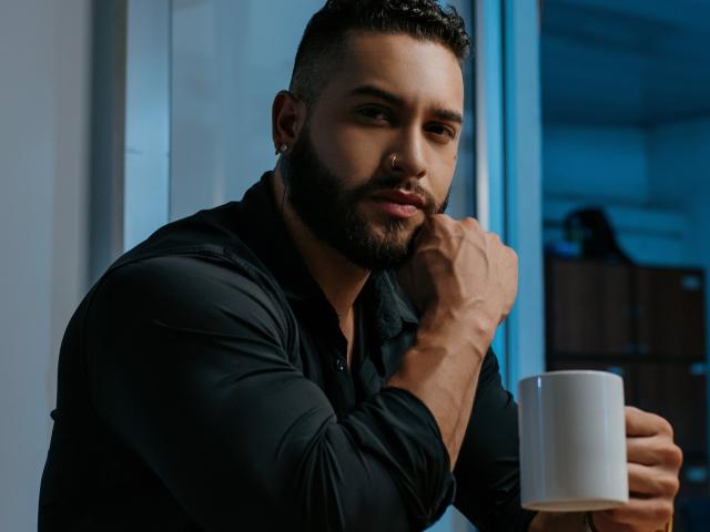 MassimoFiore with a dark beard and short hair, wearing a black shirt, holding a white mug, resting his chin on his hand, looking serious, and has a nose ring.
