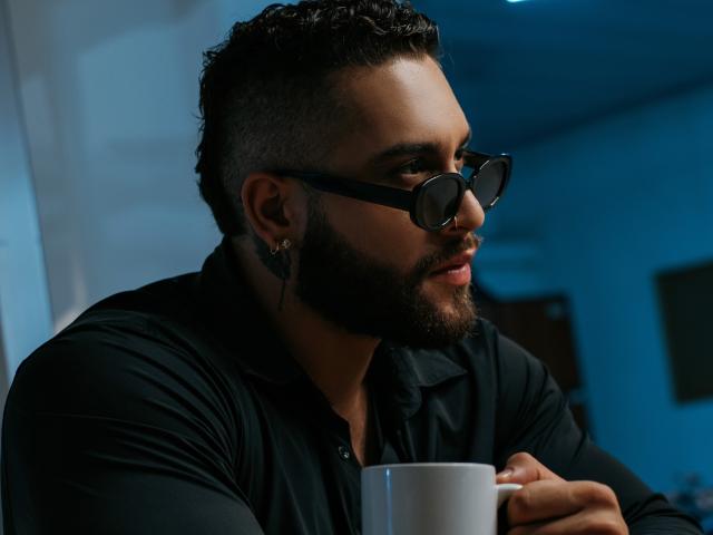 MassimoFiore with dark curly hair, beard, black glasses, black shirt, holding white mug, looking to the side, blue lighting, earring in left ear, serious expression.