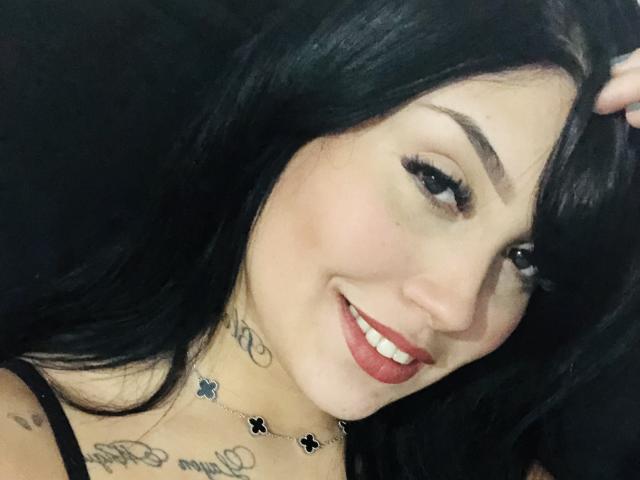 AngellHott with black hair, red lipstick, smiling, black eyeliner, tattoos on neck and chest, holding hair, wearing black spaghetti straps, necklace with small black crosses.