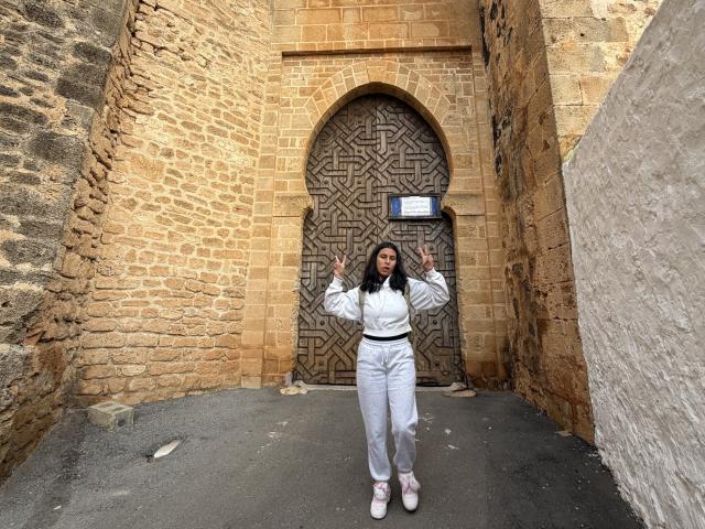 SarahPrincess stands in front of a patterned wooden door, wearing a white outfit with long sleeves and pants, and pink shoes, waving with both hands.