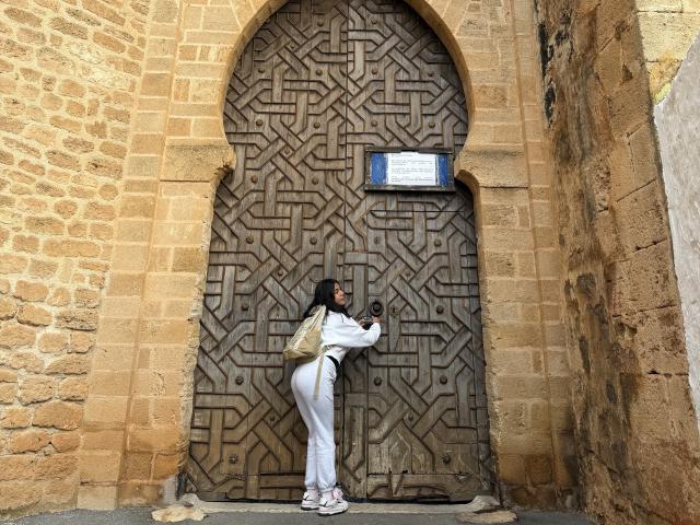 SarahPrincess stands in front of a large, intricately patterned wooden door. She faces the door, her right hand on the door's latch, and her left hand on the door's metal plate. She wears a white pantsuit, white sneakers, and a beige backpack. Her black hair is straight and falls to her shoulders. The door has a complex, geometric design with a small blue sign above it. The stone walls are textured and weathered, in varying shades of brown. The composition centers on SarahPrincess and the door, emphasizing her interaction with the patterned surface.