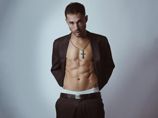 AdamsSanz, shirtless, in a brown suit, wearing a cross necklace, showing a toned, muscular, bare-chest, with hands in pockets, and black pants, against a gray background.