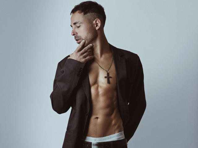 AdamsSanz with a toned, muscular, shirtless body, wearing an open brown jacket, white Calvin Klein underwear, and a black cross necklace, touching his chin with a serious expression.